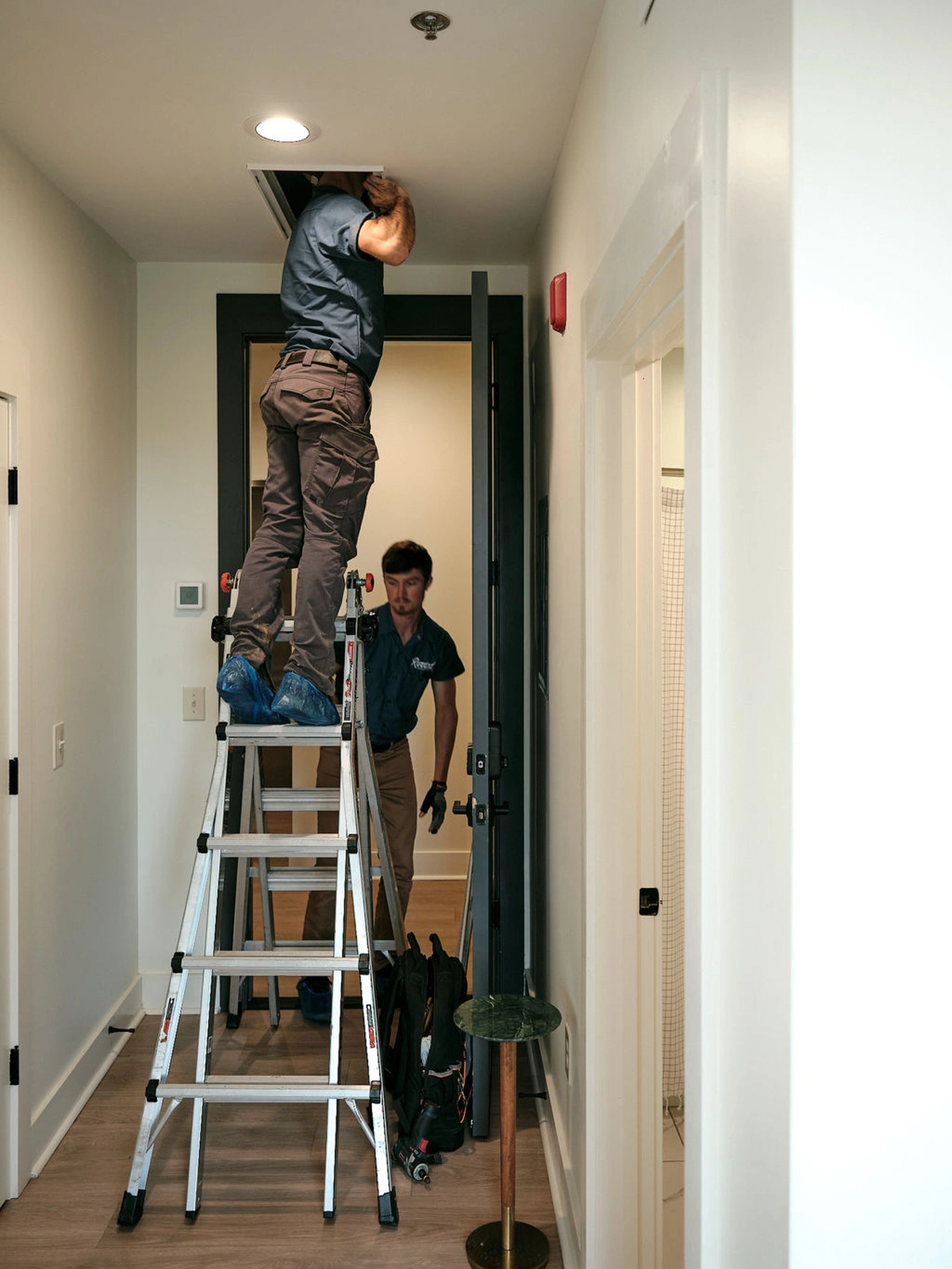 Technician on a ladder inspecting an attic access in a hallway
